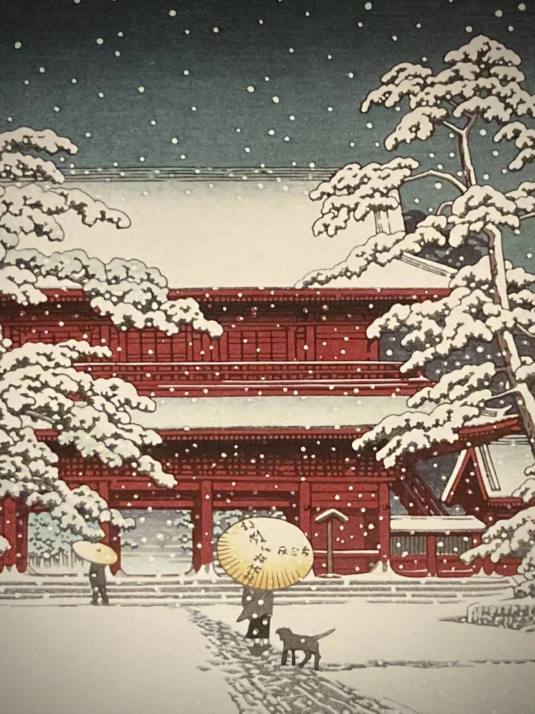 Close-up of Japanese art print by Kawase Hasui showing snow-dusted red temple gate and falling snowflakes, with figure holding umbrella on winter path.
